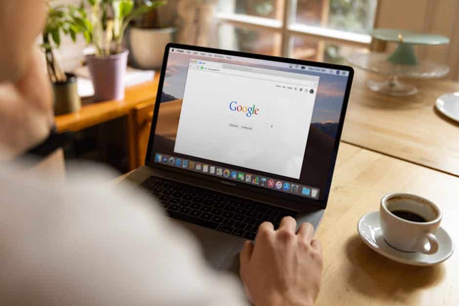 web development news - An adult using a laptop indoors, browsing Google at a wooden table with coffee.